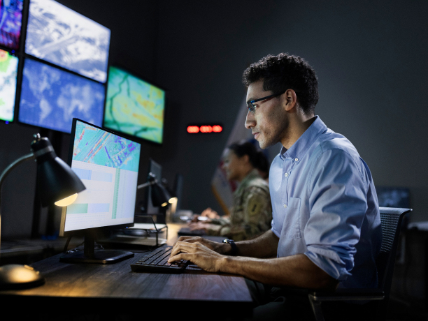 A man wearing glasses and a blue dress shirt sitting at a computer workstation next to a female Soldier in combat uniform